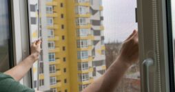 A person holding a mesh screen frame in an open window, with distant buildings visible through the net.