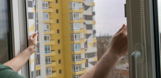 A person holding a mesh screen frame in an open window, with distant buildings visible through the net.