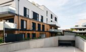 Modern apartment buildings with white walls, wood cladding, glass balconies, and underground garage ramp.