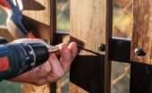 A hand holding a power drill driving a screw through a wooden plank beside metal fencing outdoors near a chain-link fence.