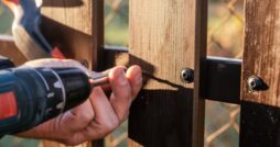 A hand holding a power drill driving a screw through a wooden plank beside metal fencing outdoors near a chain-link fence.