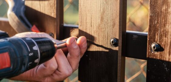 A hand holding a power drill driving a screw through a wooden plank beside metal fencing outdoors near a chain-link fence.