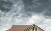 A tiled house roof under dense gray storm clouds, with a partial upper wall and dark shutters below.