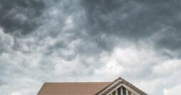 A tiled house roof under dense gray storm clouds, with a partial upper wall and dark shutters below.