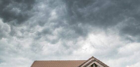 A tiled house roof under dense gray storm clouds, with a partial upper wall and dark shutters below.