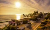 A row of oceanfront homes and palm trees lining a coastal road, with the setting sun reflecting over the ocean.