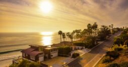 A row of oceanfront homes and palm trees lining a coastal road, with the setting sun reflecting over the ocean.