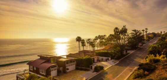 A row of oceanfront homes and palm trees lining a coastal road, with the setting sun reflecting over the ocean.