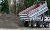 A dump truck with a raised bed unloading gravel onto a large pile near trees at an outdoor work area.