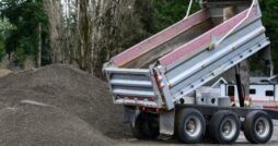 A dump truck with a raised bed unloading gravel onto a large pile near trees at an outdoor work area.