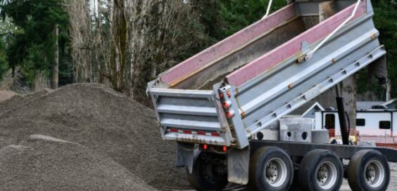 A dump truck with a raised bed unloading gravel onto a large pile near trees at an outdoor work area.