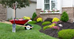 A wheelbarrow and shovel near a home’s front yard during the daytime. The yard is full of colorful flowers.