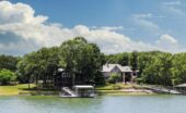 A lakeside property with trees and a private dock sits along calm water under a bright sky with scattered clouds.