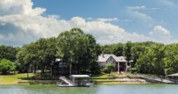 A lakeside property with trees and a private dock sits along calm water under a bright sky with scattered clouds.