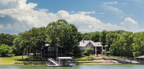 A lakeside property with trees and a private dock sits along calm water under a bright sky with scattered clouds.