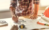 A person marking construction plans on a table with a pen, ruler, hard hat, and rolled drawings nearby.