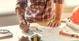 A person marking construction plans on a table with a pen, ruler, hard hat, and rolled drawings nearby.