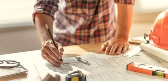 A person marking construction plans on a table with a pen, ruler, hard hat, and rolled drawings nearby.