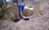 A blue capped well pipe and curved white conduit sit in excavated soil beside a wooden post and dry grass.