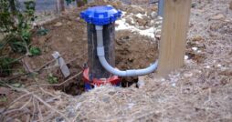 A blue capped well pipe and curved white conduit sit in excavated soil beside a wooden post and dry grass.