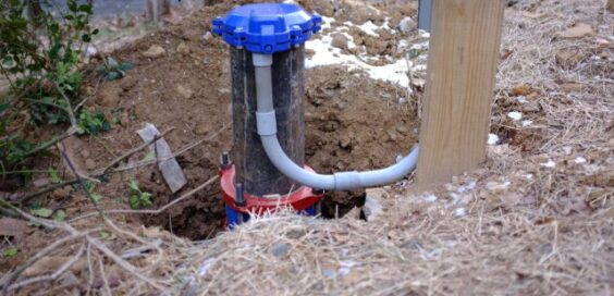 A blue capped well pipe and curved white conduit sit in excavated soil beside a wooden post and dry grass.