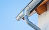 A metal gutter and downspout attached to a house roof edge, with a light wall and clear blue sky behind it.