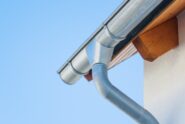 A metal gutter and downspout attached to a house roof edge, with a light wall and clear blue sky behind it.