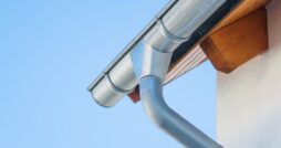 A metal gutter and downspout attached to a house roof edge, with a light wall and clear blue sky behind it.