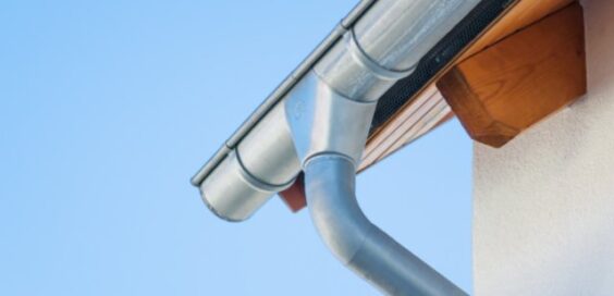 A metal gutter and downspout attached to a house roof edge, with a light wall and clear blue sky behind it.