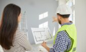 A builder and a homeowner review a floor plan while pointing toward an unfinished room during a home construction project.