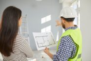A builder and a homeowner review a floor plan while pointing toward an unfinished room during a home construction project.