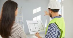 A builder and a homeowner review a floor plan while pointing toward an unfinished room during a home construction project.