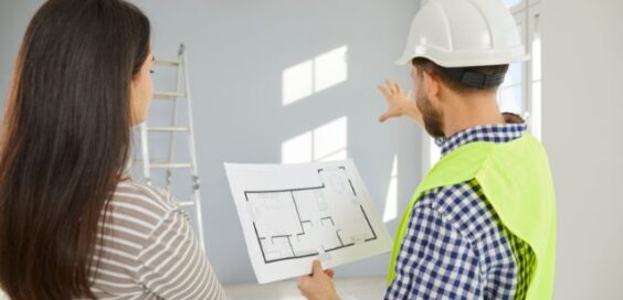 A builder and a homeowner review a floor plan while pointing toward an unfinished room during a home construction project.