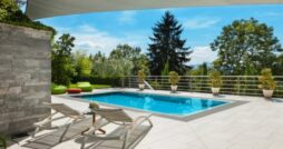 Rectangular backyard pool surrounded by a tiled patio, lounge chairs, potted plants, and lush green trees.