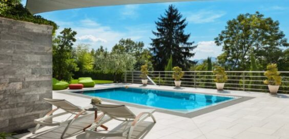 Rectangular backyard pool surrounded by a tiled patio, lounge chairs, potted plants, and lush green trees.