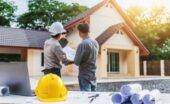 A person in a hard hat points near a house while another person holds plans at a table in the foreground.