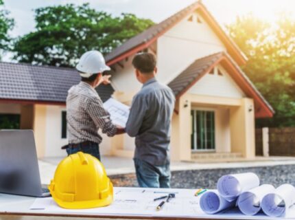 A person in a hard hat points near a house while another person holds plans at a table in the foreground.