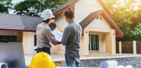 A person in a hard hat points near a house while another person holds plans at a table in the foreground.