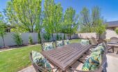 A backyard space with a patio, outdoor dining table, cushioned chairs, trees, and a white privacy fence.