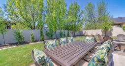 A backyard space with a patio, outdoor dining table, cushioned chairs, trees, and a white privacy fence.