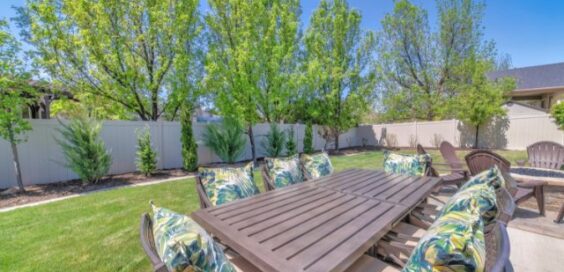 A backyard space with a patio, outdoor dining table, cushioned chairs, trees, and a white privacy fence.