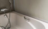 Light shining on a rectangular acrylic bathtub in a residential bathroom. A shower head attachment is on the wall.