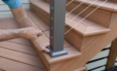 A worker tightens a cable railing fitting on a dark metal post beside the wooden stairs of a backyard deck.