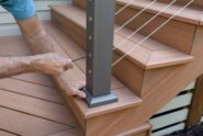 A worker tightens a cable railing fitting on a dark metal post beside the wooden stairs of a backyard deck.