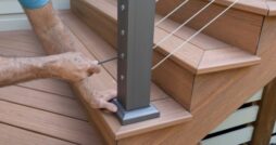 A worker tightens a cable railing fitting on a dark metal post beside the wooden stairs of a backyard deck.