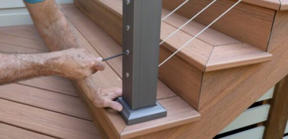 A worker tightens a cable railing fitting on a dark metal post beside the wooden stairs of a backyard deck.