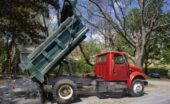 A dump truck with a green bed and red cab dumps aggregate in the front yard of a residential property.