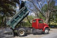 A dump truck with a green bed and red cab dumps aggregate in the front yard of a residential property.