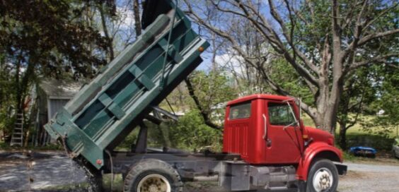 A dump truck with a green bed and red cab dumps aggregate in the front yard of a residential property.