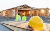 Two people wearing yellow hard hats and yellow safety vests are standing next to a home construction site.